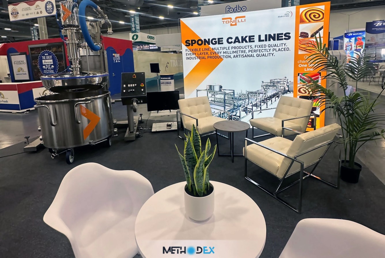 Methodex-built Tonelli booth with cream lounge chairs, a round table, stainless steel baking equipment, and a backlit Sponge Cake Lines graphic