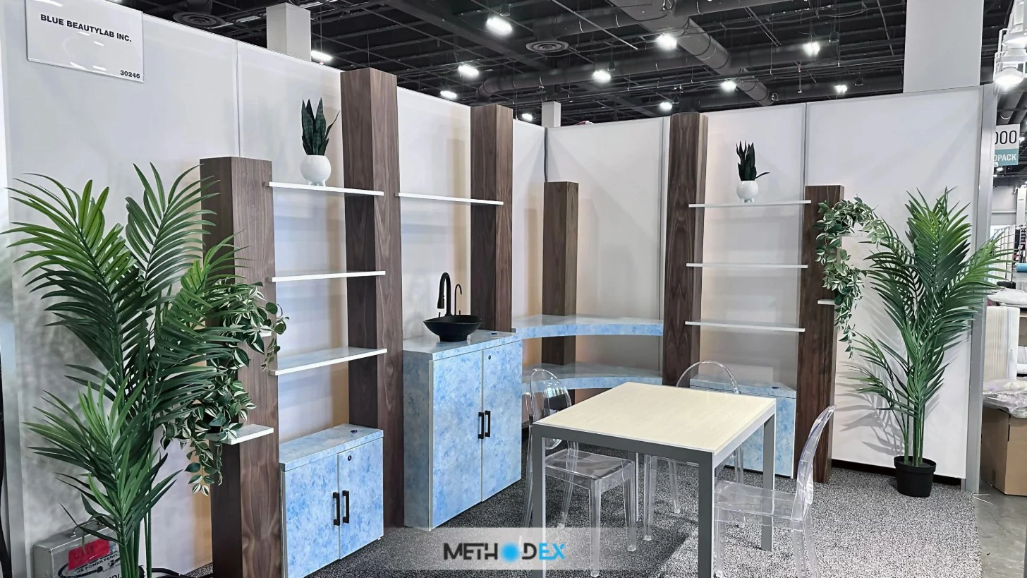 Blue Beauty Lab booth with white walls, wood-look posts with white shelves, blue cabinets with a black vessel sink, a small table with clear chairs, and potted plants