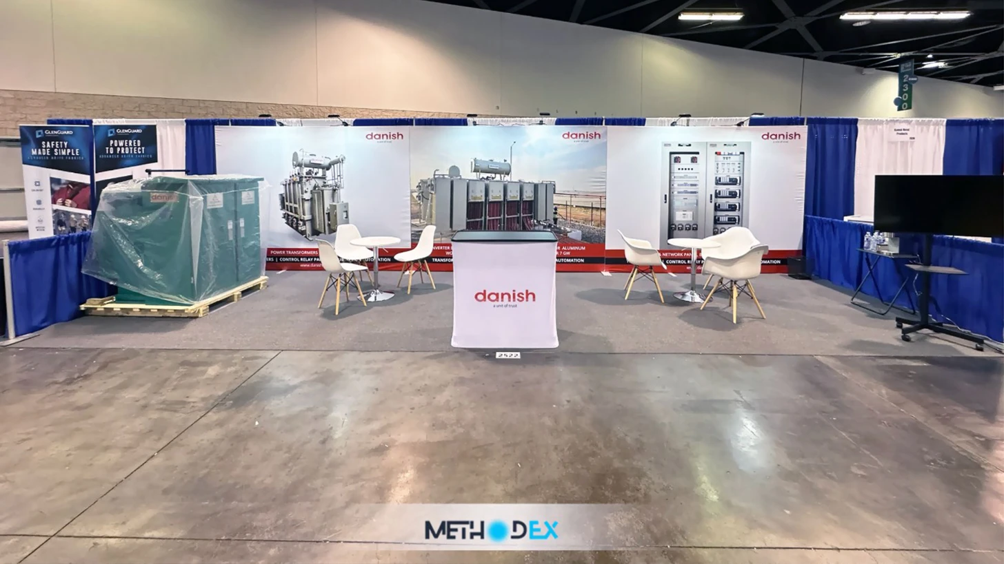 Inline booth with a printed backwall showing transformers and electrical panels, a branded counter, white chairs and tables, a plastic-wrapped green unit on a pallet, and a monitor on a stand