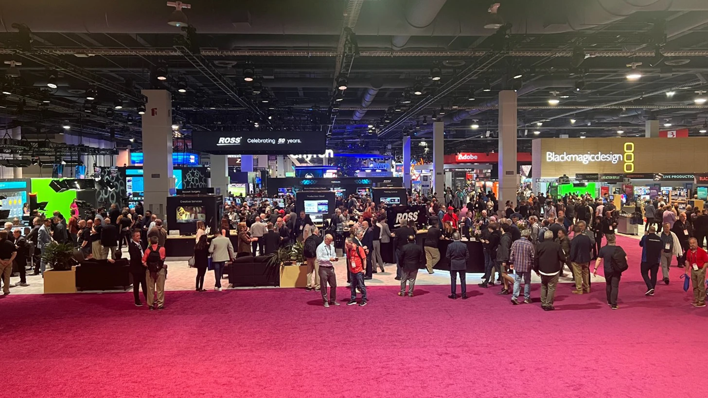 Crowded exhibit floor with magenta carpet, black booth structures, and overhead signs for Ross, Adobe, and Blackmagic Design