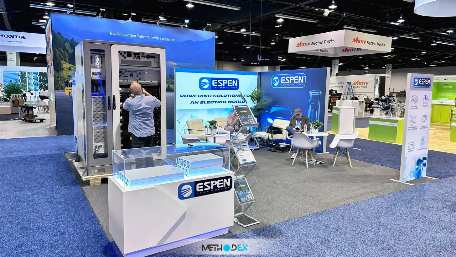 Espen Technology island booth with backlit blue graphic walls, a lit white counter, a glass product display case, and a lounge area