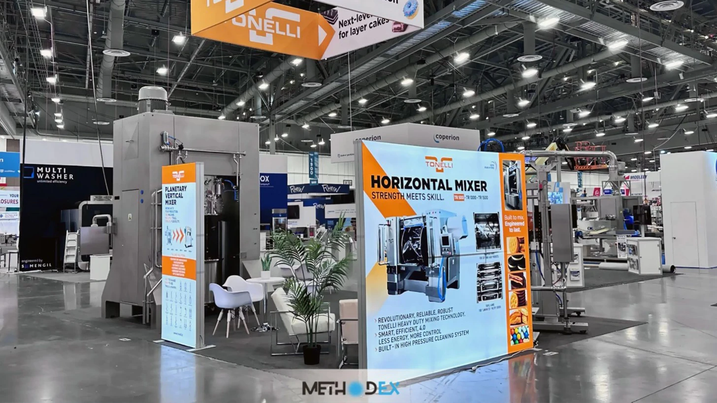 Tonelli booth with an orange and white hanging sign, a large stainless steel mixer, backlit product graphics, a potted palm, white chairs, and a white bench