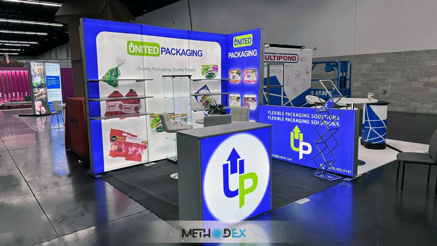 United Packaging booth with backlit blue and white walls, shelves of produce packaging, a glass display case, white chairs, and a lit counter with the UP logo